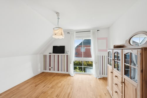 Helles Kinderzimmer mit Holzboden, Dachschräge, großem Fenster, einem TV auf einer Kommode und einem Holzschrank.