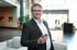 Ein Mann im Anzug mit Brille hält ein Smartphone in einem modernen Innenraum.