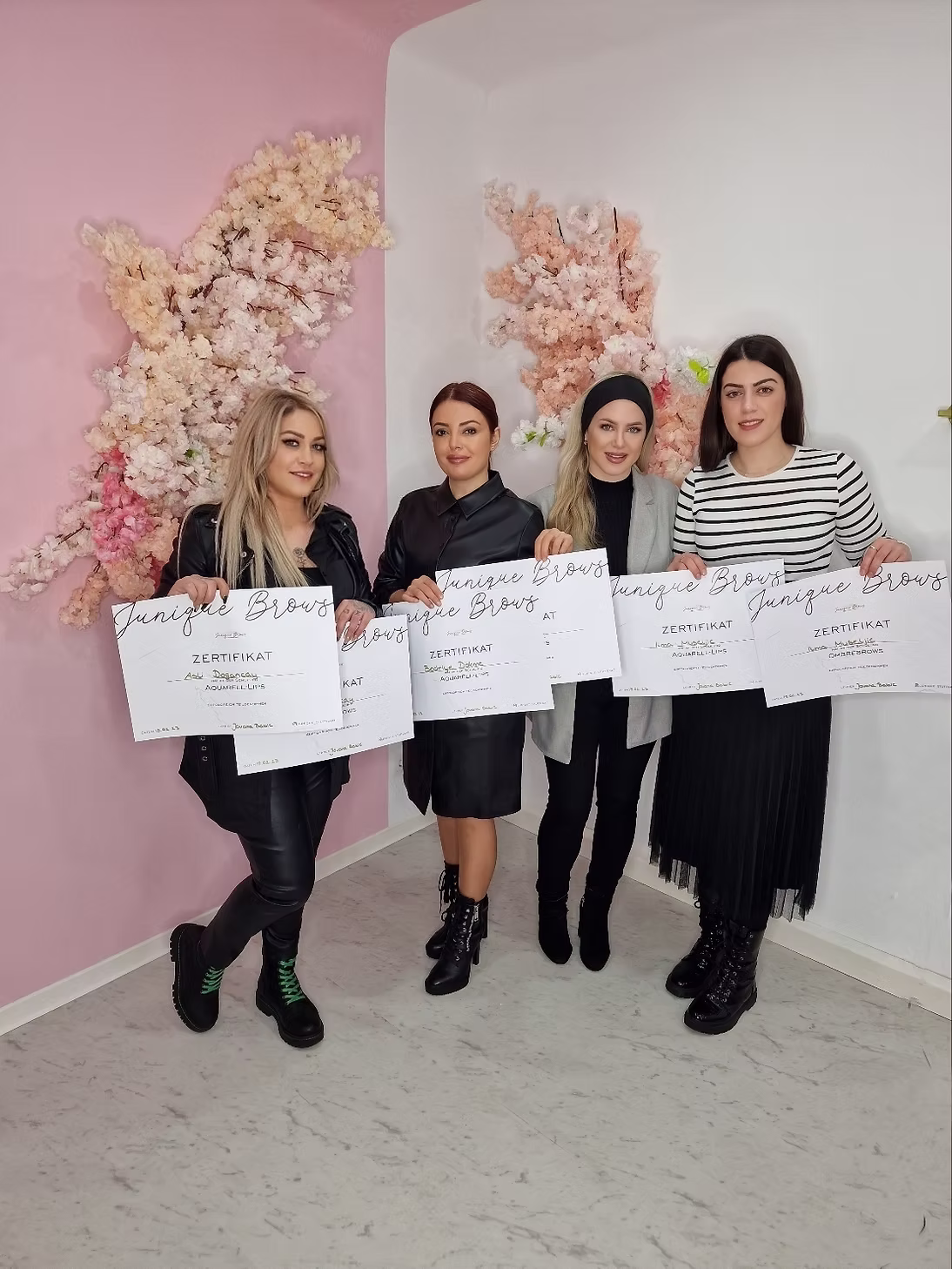 Vier Frauen halten Zertifikate mit der Aufschrift „Junique Brows“ vor einer rosa Wand mit floralen Dekorationen.