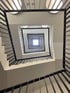 Blick nach oben in ein quadratisches Treppenhaus mit schwarzem Geländer, das sich spiralförmig nach oben windet.