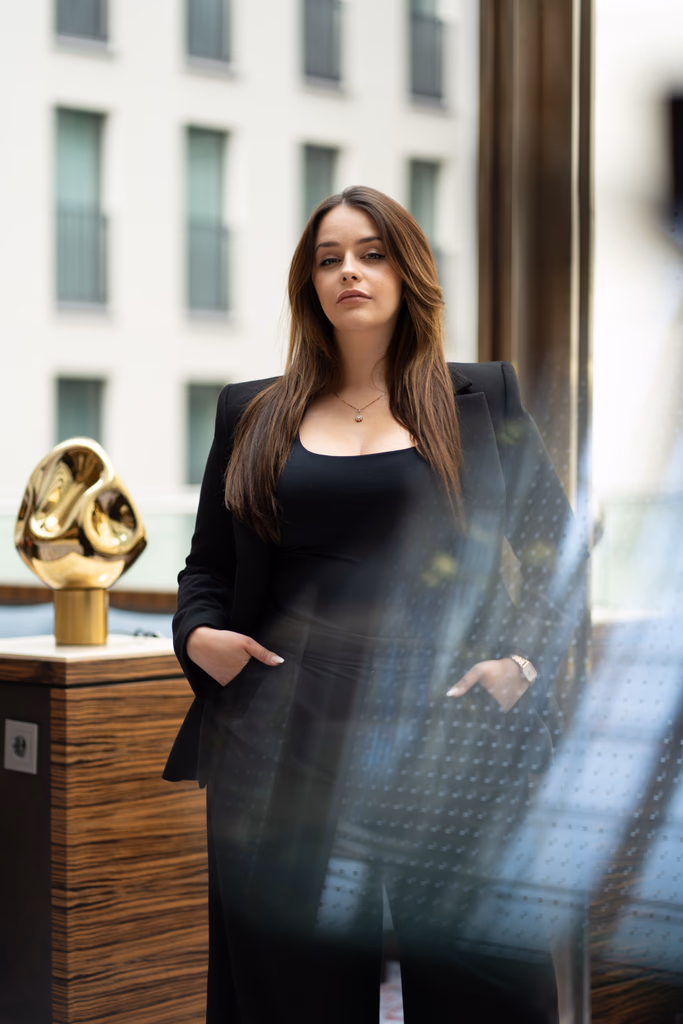 Junge Frau mit langen braunen Haaren und schwarzem Blazer steht vor einem modernen Gebäude mit einem goldenen Objekt.