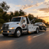 Towing Deltona tow truck transporting a disabled sedan along a highway at sunset