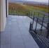 Eine moderne Terrasse mit hellgrauen Fliesen und Glasgeländer, die zu einem Weinberg mit kahlen Reben führt.