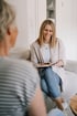 Eine lächelnde Frau sitzt auf einem Sofa, hält ein Notizbuch und spricht mit einer anderen Person.