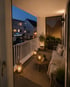Beleuchteter Balkon am Abend mit Sofa, Couchtisch, Teppich, Pflanzen und Laternen vor Stadtkulisse.