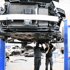 Zwei Mechaniker arbeiten unter einem auf einer Hebebühne angehobenen Auto in einer Werkstatt.