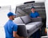 Zwei Umzugshelfer in blauen Uniformen laden ein graues Sofa in einen weißen Lieferwagen.