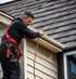 Ein Handwerker mit schwarzer Kleidung und rotem Sicherheitsgurt reinigt eine Dachrinne an einem beige verkleideten Haus mit dunklem Schindeldach.