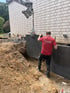 Ein Bauarbeiter von GartenGlück setzt mithilfe eines Krans eine große dunkle Steinplatte in einen Graben vor einer Hauswand.