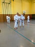Zwei Männer in weißen Karate-Anzügen trainieren in einer Turnhalle, weitere Schüler stehen im Hintergrund.