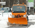 Orangefarbener Schneepflug räumt eine verschneite Straße von vorne.