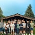 Eine Gruppe von 23 jungen Erwachsenen steht lächelnd vor einer Holzhütte mit einem Baum im Hintergrund.