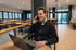 Ein lächelnder Mann telefoniert mit einem Smartphone und arbeitet an einem Laptop in einem Café.