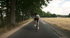 Ein Mann fährt auf einem Rennrad eine asphaltierte Straße entlang, gesäumt von hohen Bäumen und einem Feld.