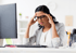 Eine Frau mit dunklen Haaren und Brille sitzt an einem Schreibtisch und hält sich die Schläfen, ihr Blick ist gesenkt und wirkt angespannt. Ein Computerbildschirm ist unscharf im Hintergrund zu sehen.