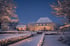 Beleuchtetes weißes Haus in einer verschneiten Gartenlandschaft bei Dämmerung mit lichtergeschmückten Hecken und Bäumen.