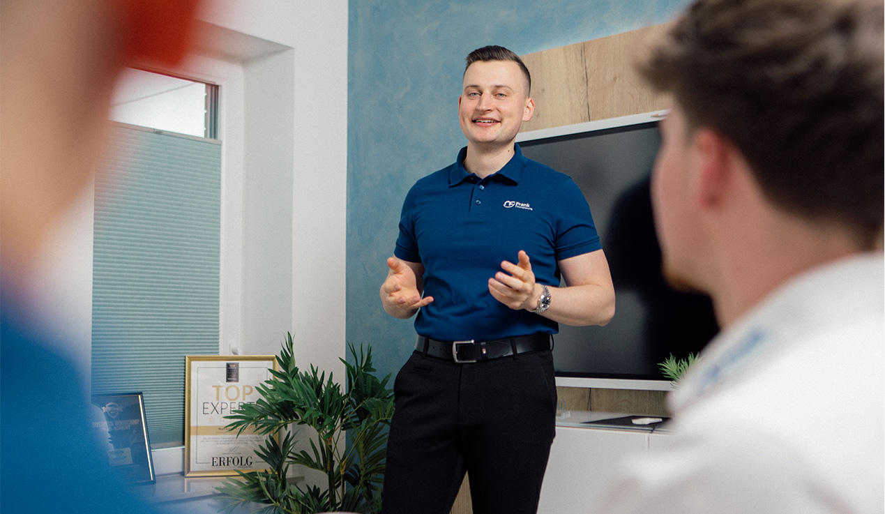 Ein lächelnder Mann im blauen Poloshirt hält einen Vortrag vor einer Gruppe in einem modernen Büro.
