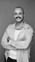 Ein lächelnder Mann mit Schnurrbart, Nasenring und Glatze, der eine gestreifte Jacke trägt.