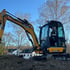 Gelber JCB 8029 CTS Minibagger mit Fahrer arbeitet auf einem Erdhaufen vor Bäumen und Wohnhäusern.