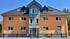Orangefarbenes Mehrfamilienhaus mit grauem Mittelteil, dunklem Dach und schwarzem Metallzaun.