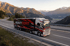 Schwarzer Heavy Cont Austria LKW fährt auf einer Bergstraße mit verschneiten Gipfeln und einem Tal im Hintergrund.