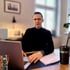 Mann mit Brille und schwarzem Rollkragenpullover sitzt am Schreibtisch vor einem Laptop.
