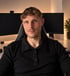 Ein junger Mann mit blondem Haar sitzt in einem Gaming-Stuhl vor zwei Monitoren an einem Schreibtisch.