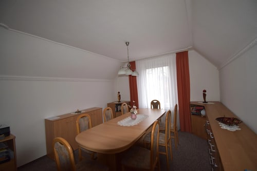 Esszimmer mit ovalem Holztisch, sechs Stühlen, Sideboards und Fenster mit weißen Vorhängen und roten Gardinen.