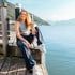 Frau mit lockigem Haar sitzt lächelnd auf einem Holzsteg am See mit Bergen im Hintergrund.