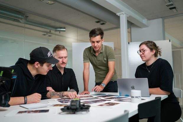 Vier Personen sitzen und stehen um einen Tisch, prüfen Fotos und schauen auf einen Laptop.