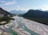 Ein türkisblauer Fluss schlängelt sich durch ein breites Kiesbett, umgeben von grünen Wäldern und Bergen unter blauem Himmel.