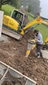 Ein gelber Wacker Neuson Bagger und eine Person mit einem Vibrationsstampfer arbeiten auf einer Erdbaustelle im Freien.