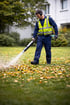 Ein Landschaftsgärtner in blauer Uniform und gelber Warnweste benutzt einen Laubbläser, um Herbstlaub auf einer Wiese zu entfernen.