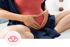 A pregnant person sits with hands forming a heart shape over their belly, wearing a red top and blue fabric.