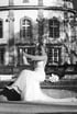 Eine Braut im weißen Hochzeitskleid öffnet lachend eine Sektflasche vor einem historischen Gebäude in Schwarz-Weiß.