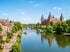 Schloss Johannisburg, ein Fluss mit Booten, eine Uferpromenade mit Menschen und eine Brücke unter blauem Himmel.