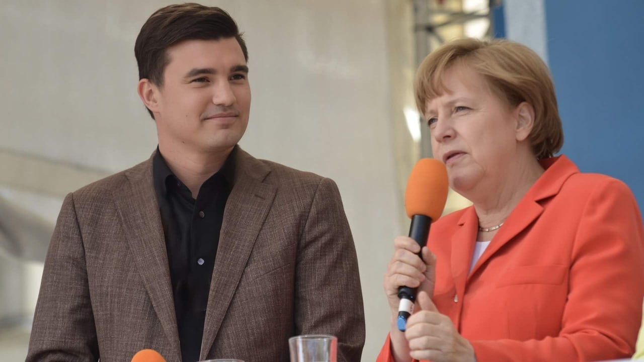Tim Christopher Gasse neben Bundeskanzlerin a. D. Dr. Angela Merkel, die in ein Mikrofon spricht