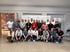 Eine Gruppe von 26 Personen, meist Männer, posiert in einem modernen Raum vor einer Wand mit Firmenlogos.
