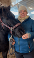 Eine Frau in blauer Jacke, beigefarbener Mütze und Schal steht neben einem schwarzen Pferd, das ein rotes Halfter trägt.