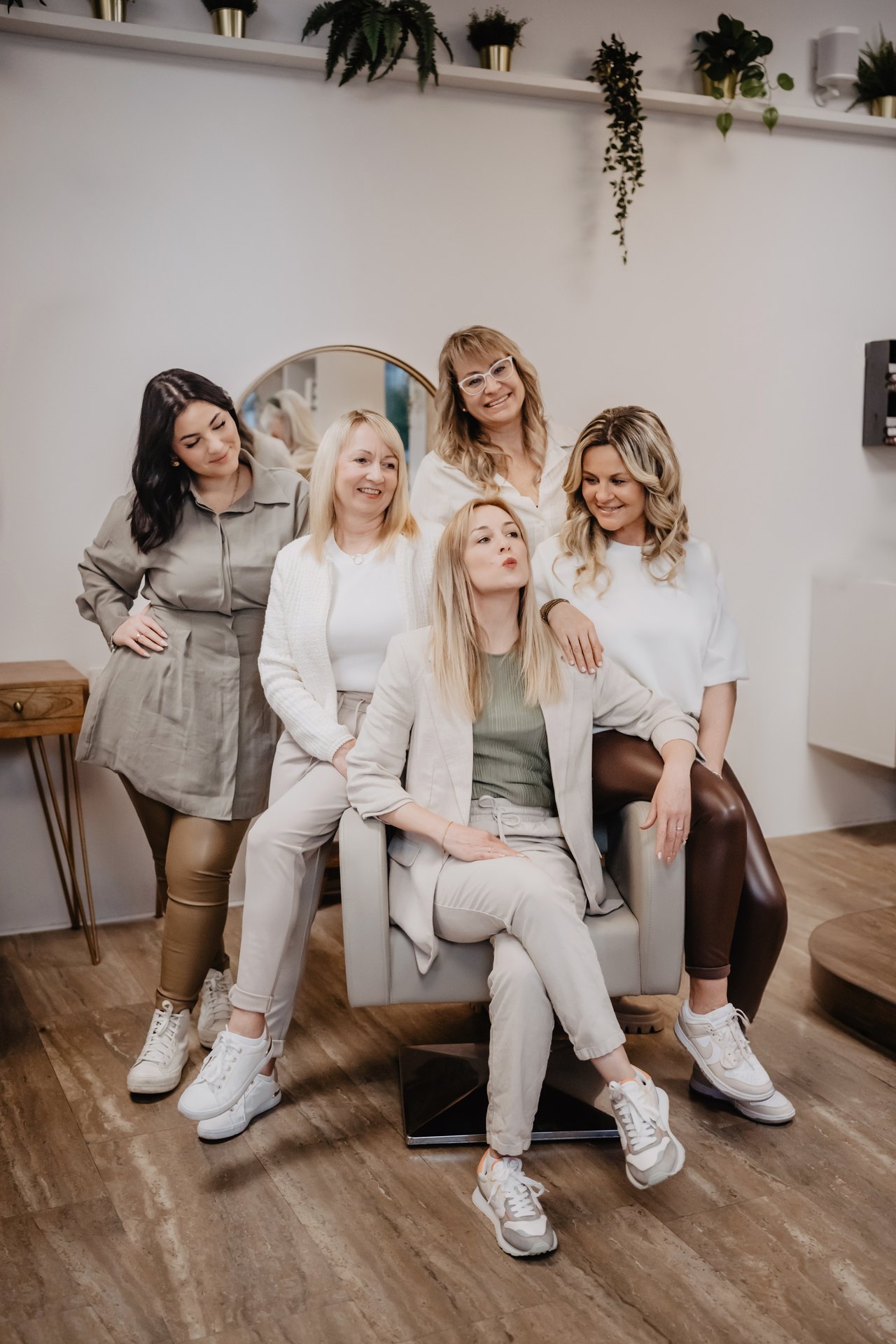 Fünf lächelnde Frauen in heller Kleidung posieren gemeinsam in einem modern eingerichteten Friseursalon mit Zimmerpflanzen.