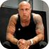 Bald man with tattooed arms, wearing a black tank top and silver chain, sits with hands clasped, looking forward.