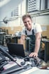 Ein lächelnder Mechaniker in Arbeitskleidung beugt sich über den offenen Motor eines Autos und arbeitet an einem Laptop.