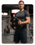 Muskulöser Mann in grauem T-Shirt und blauen Shorts, der eine schwarze Hantelscheibe im Fitnessstudio hält.