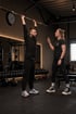 Ein Mann führt im Fitnessstudio Überkopfdrücken mit einer Langhantel aus, während eine Trainerin Anweisungen gibt.