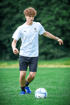 Junger Mann mit lockigem Haar in weißem T-Shirt und schwarzer Hose dribbelt einen Fußball auf grünem Gras.