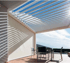 Eine modern designte Pergola mit Lamellendach über einer Terrasse mit Möbeln und einem Sonnenschutz.