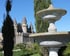 Steinerner Springbrunnen vor einem Schloss mit spitzen Türmen, umgeben von grünen Zypressen und Büschen.