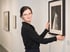 Frau mit Brille und schwarzer Bluse hängt ein gerahmtes Schwarz-Weiß-Foto an einer weißen Galeriewand auf.