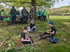 Kinder sitzen im Gras und schnitzen Stöcke, während eine Wandergruppe im Hintergrund unter einem Baum rastet.