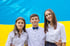 Drei lächelnde Jugendliche in weißen Hemden stehen vor einer großen blau-gelben Flagge der Ukraine.